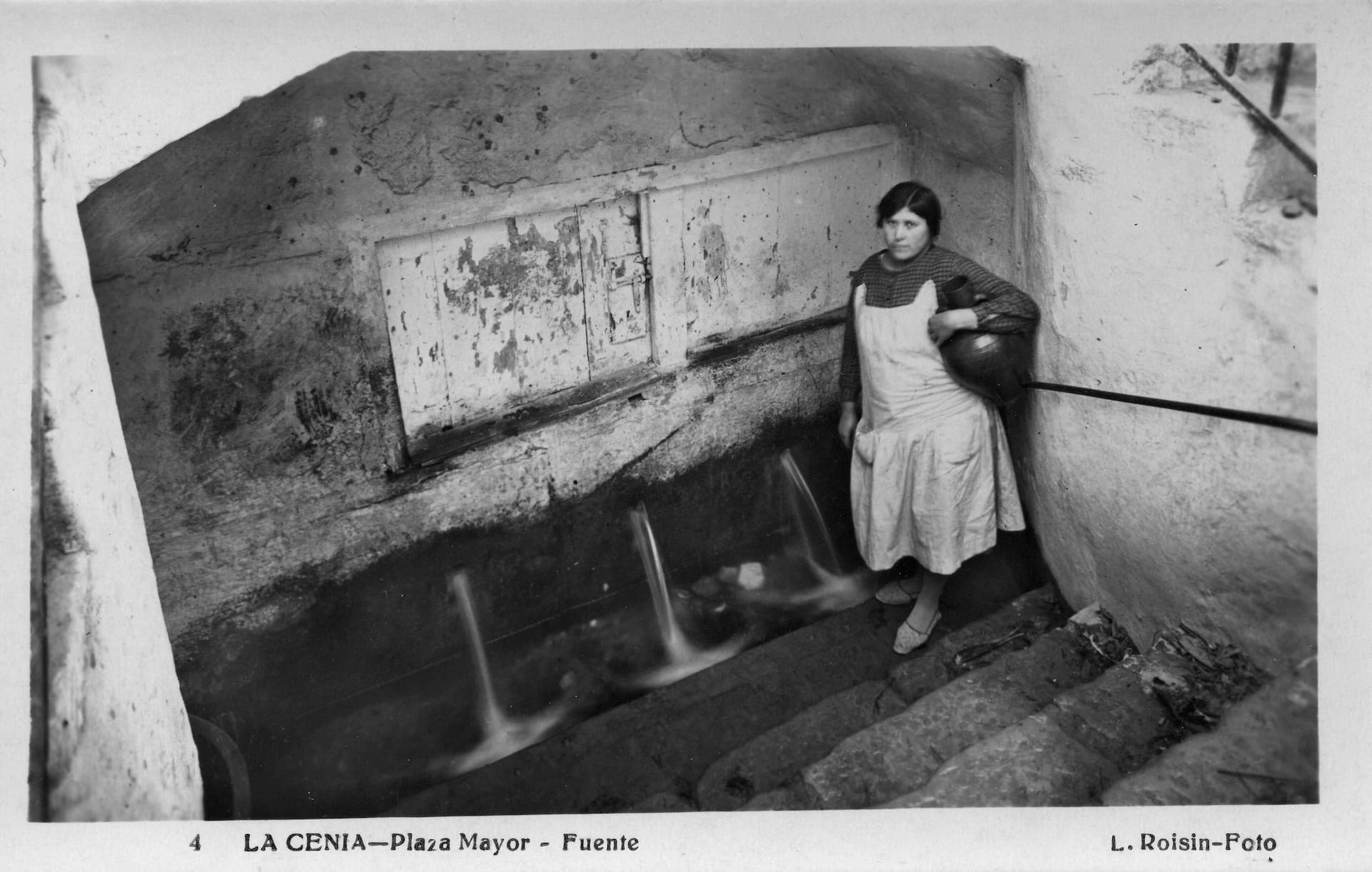 Dona amb un c&agrave;nter baix del bra&ccedil; que va a buscar aigua a la Font Vella de la pla&ccedil;a. Anys 30 segle XX. Font: Fons Lucien Roisin &ndash; Centre d&rsquo;Estudis Seniencs.